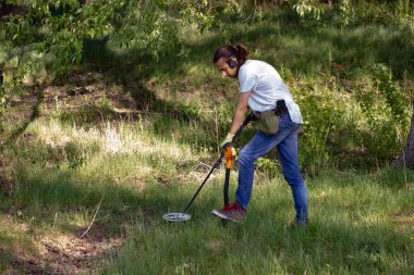 a young man in jeans, a black sweatshirt and wireless headphones is searching the ground in search of treasure using a pinpointer. nearby lies a wireless metal detector and a shovel stuck into the gro