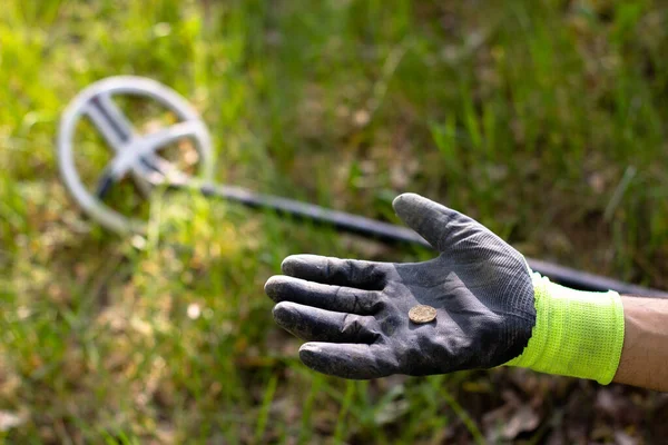 a coin found with a wireless metal detector lies on the palm of a glove. in the background, the metal detector coil lies in the center of the frame. among the young grass