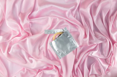 A piece of a condom sticks out of a foil package against a pink silk sheet. Safe sex and reproductive health concept.