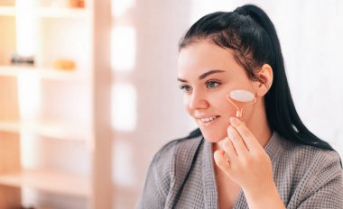 Jade Gouache ile yüz gerdirme ameliyatı için masaj. Yüz masajı silindiri kullanan Avrupalı görünümlü güzel bir kadın. Evde cilt bakımının güzelliği