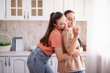 İki genç kız mutfakta pencerenin yanında duruyor ve nazikçe sarılıyorlar. LGBT lezbiyen çifti mutluluk anlarını sever
