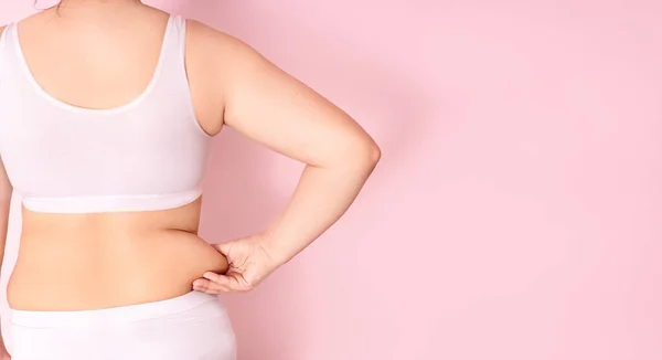 Tlustá dívka drží ruku s přebytečným tukem na břiše na růžovém pozadí. koncept ženského životního stylu s dietou ke snížení břicha. zotavení po porodu. místo pro text — Stock fotografie