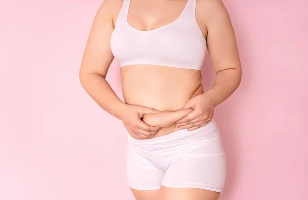 Tlustá dívka drží ruku s přebytečným tukem na břiše na růžovém pozadí. koncept ženského životního stylu s dietou ke snížení břicha. zotavení po porodu. místo pro text — Stock fotografie