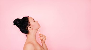 Kafkasyalı bir kadının, boynuna yeşim bir silindirle masaj yaparken görüntüsü. Tatmin olmuş ve güzel bir esmer bayanın tadını çıkarıyor. Vücut ve cilt bakımı. Pembe arka planda izole edilmiş. Stüdyo çekimi