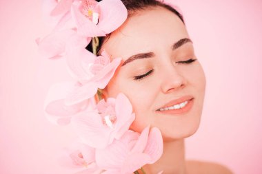 Gözleri kapalı, genç ve güzel bir kadın. Doğal makyajı ve pembe arka planda muhteşem pembe bir orkidesi olan taze bir teni var.