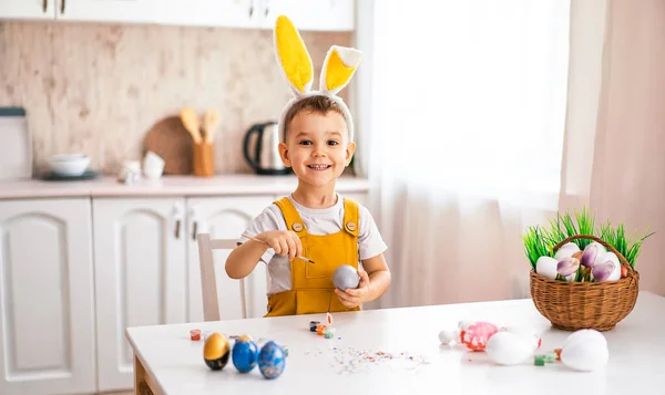 一个相貌欧洲的英俊小男孩笑着从桌上的复活节彩蛋上画了出来 头上戴着一只黄兔耳朵 — 图库照片