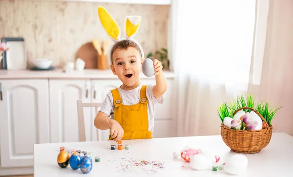 一个相貌欧洲的英俊小男孩笑着从桌上的复活节彩蛋上画了出来 头上戴着一只黄兔耳朵 — 图库照片