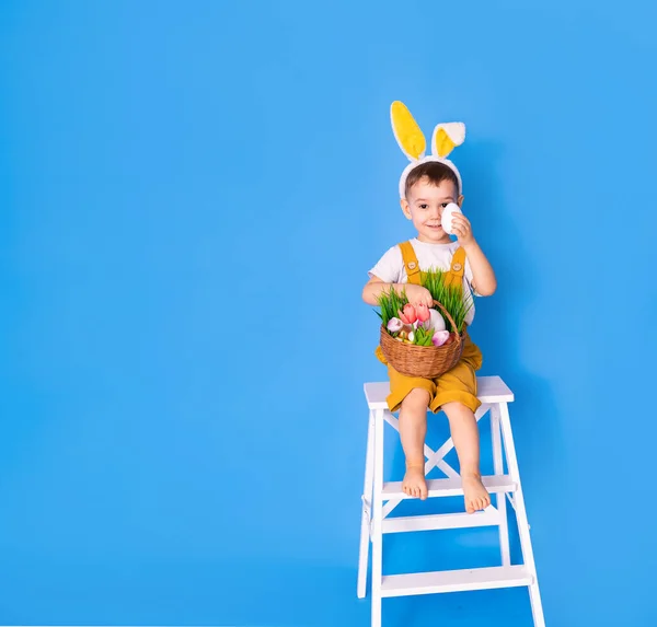 一个穿着黄色连衣裙 头上戴着小兔子耳朵的可爱的小男孩 一边拿着复活节篮子一边笑 — 图库照片