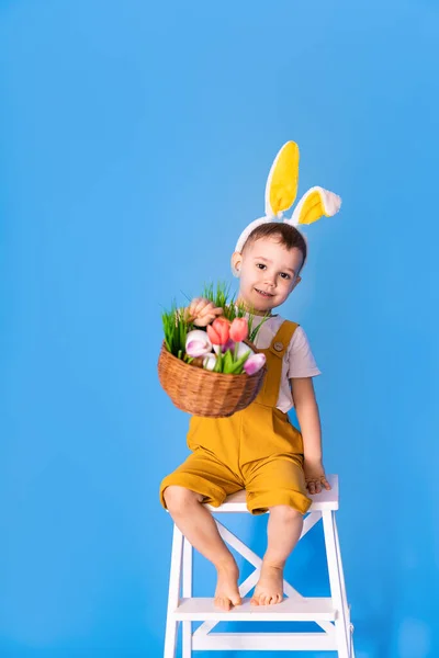 一个穿着黄色连衣裙 头上戴着小兔子耳朵的可爱的小男孩 一边拿着复活节篮子一边笑 — 图库照片