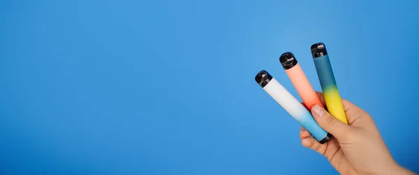 Disposable three electronic cigarettes in a woman's hand close-up on a blue background. The concept of modern smoking, vaping and nicotine. Space for text.