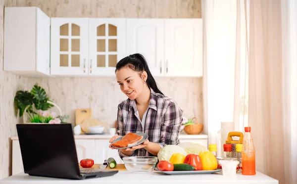 Eine fröhliche junge Frau kocht in der Küche mit einem Online-Publikum. Das Mädchen zeigt den roten Fisch ihrem Online-Publikum. Online kochen. — Stockfoto