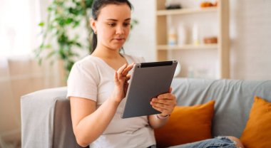 Gülümseyen genç bir kız evdeki dijital tablette sanal bir sınıfta internetten ders çalışıyor. Evde kanepede tabletli bir kız. seçici odak