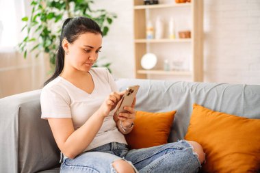 Esmer bir kız evdeki sosyal ağları kontrol ediyor. Avrupa görünümlü gülümseyen genç bir kız, telefon kullanıyor, oyun oynuyor, online alışveriş yapıyor, kanepeye sipariş veriyor.