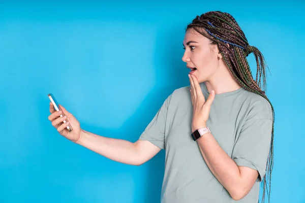 a young beautiful woman experiencing happiness or unexpected shock and looking at a smartphone on a blue background can be used for advertising or presentation of a product concept