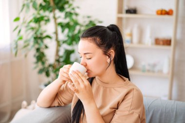 A happy young woman drinks a cup of tea on an autumn morning. A dreamy girl is sitting in the living room with a cup of hot coffee. A beautiful woman at home