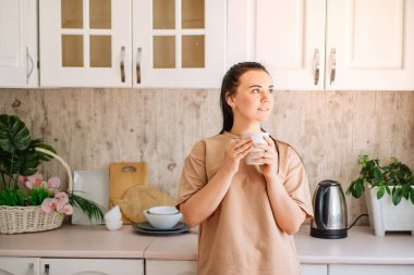 A young happy brunette woman drinks coffee in the kitchen in the morning