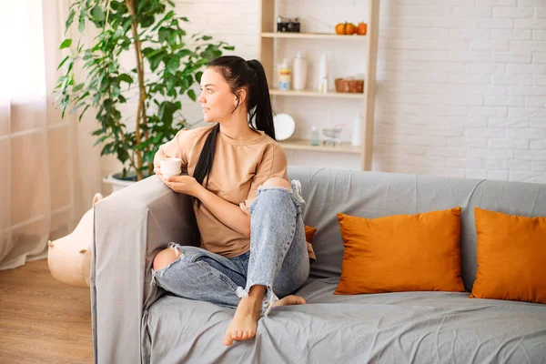 A happy young woman drinks a cup of tea on an autumn morning. A dreamy girl is sitting in the living room with a cup of hot coffee. A beautiful woman at home