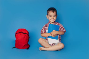 Neşeli, gülümseyen küçük bir esmer elinde bir sırt çantası ve defterlerle oturmuş mavi bir duvarın arkasında eğleniyor. Okul konsepti. Okula geri dön..