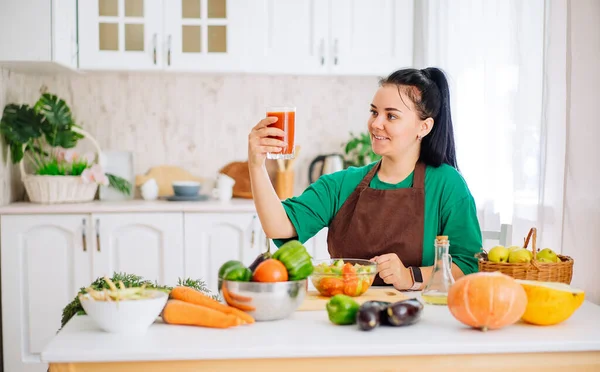 A young brunette woman in an apron is sitting at a table with fresh vegetables, looking at a glass of freshly squeezed homemade juice from fresh vegetables in a white kitchen