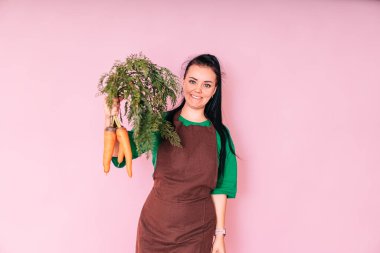 a beautiful farmer girl holds a carrot on a pink background. the concept of harvesting.
