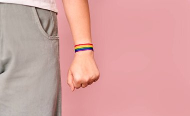 Kolunda LGBT bayrağı renginde bir bilezik takan genç bir kadının eli, üzerinde LGBT bileziği olan bir yumruğa yapışmış, pembe arka planda işaretlenmiş.