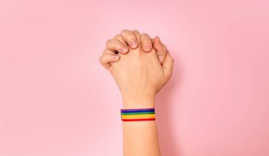 İki Asyalı, pembe arka planda bileklerinde gökkuşağı deseni olan bir bileklikle el ele tutuşuyorlar. Igbt, eşcinsel ilişkiler ve homoseksüeller..