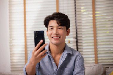 Asyalı adam evde akıllı telefonuyla görüntülü arama yapıyor. Telefon ekranında el sallıyor. Konferans toplantısı uygulaması kullanılıyor.