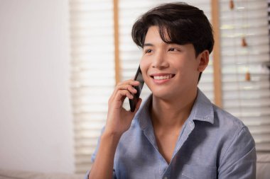 Gülümseyen Asyalı genç işadamı evde telefonla konuşuyor, iş görüşmesi yapıyor ve kurumsal mobil tartışmanın tadını çıkarıyor.