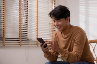 Evdeki kanepede oturan Asyalı bir adam akıllı telefonuyla çalışıyor. Adam sosyal medya uygulamalarını kullanırken çalışıyor. Evden çalışma kavramı..