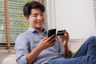 Asyalı adam internet üzerinden ödeme yapıyor. Evdeki kanepede, internetten alışveriş yapan biri kredi kartı ve akıllı telefon kullanıyor. İnternet alışverişi kavramı.