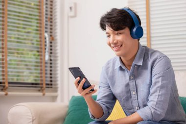 Evdeki kanepede oturan Asyalı bir adam akıllı telefonuyla çalışıyor. Adam sosyal medya uygulamalarını kullanırken çalışıyor. Evden çalışma kavramı..