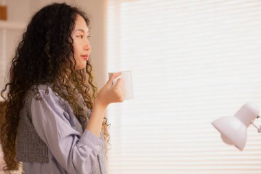 Asain kadın sabah kahvesini yudumluyor ve beyaz bir fincan kahveyle uyanıyor. Hafta sonları dinleniyorum.