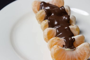 Mandalina beyaz bir tabakta çikolatayla dilimler. Erimiş çikolatayla kaplı soyulmuş mandalina. Meyveli tatlı.