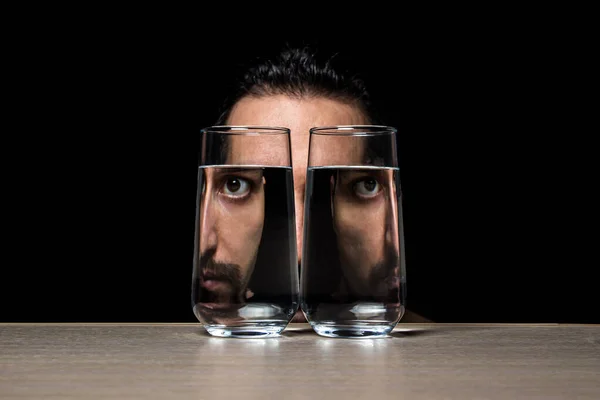 Bir adam siyah arka planda iki bardak suyun içinden bakar. Sudaki bozulma. yaratıcı portre