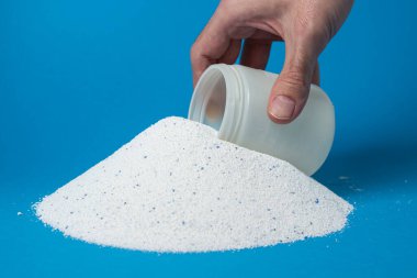 Mavi arka planda çamaşır deterjanı. Bir el ölçü bardağında çamaşır tozu topluyor. Ev kimyasalları.