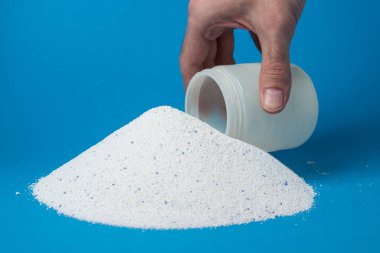 Mavi arka planda çamaşır deterjanı. Bir el ölçü bardağında çamaşır tozu topluyor. Ev kimyasalları.