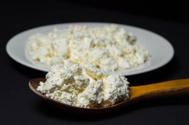 Siyah arka planda bir tabak süzme peynir. Sağlıklı ve lezzetli süt ürünleri. Ahşap kaşık dolusu süzme peynir.