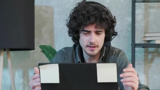 Mladý kudrnatý muž pomocí notebooku se sluchátky mluvit on-line. — Stock video