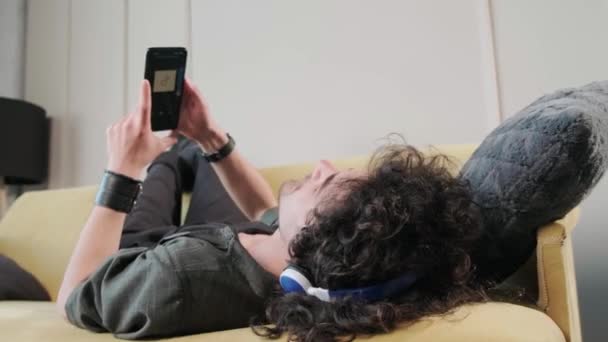 Chico de pelo rizado en el sofá escuchando música con auriculares, divirtiéndose. — Vídeos de Stock