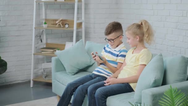 笑顔の妹と一緒に携帯アプリを楽しんでいる幸せな弟 ソファの上に一緒に座ってスマートフォンを使用して興奮した双子の子供 — ストック動画