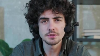 Kıvırcık saçlı genç adam kameraya bakıyor ve kulaklıkla konuşuyor..