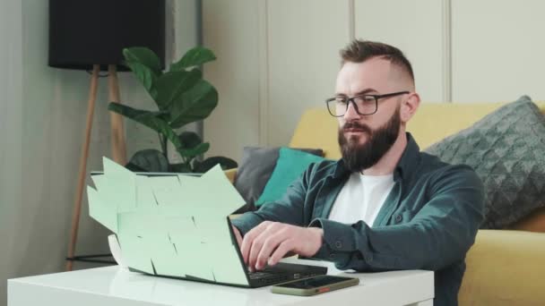 Homem trabalhando no computador com um monte de notas de lembrete, sentado perto do sofá. — Vídeo de Stock