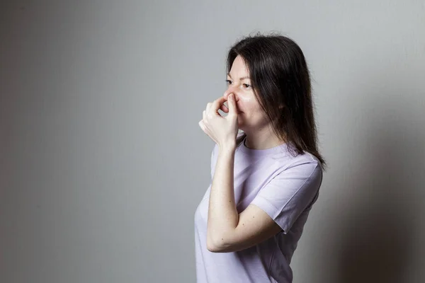 What a smell. Close up of young unhappy beautiful woman with long hair in casual t-shirt squeezing nose with fingers, disgust expression, feeling bad about bad smell on street