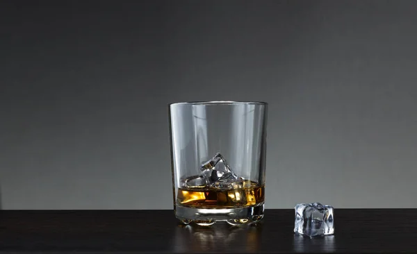 glass of whiskey with ice on a wooden table. A glass of whiskey and a piece of ice on the table.