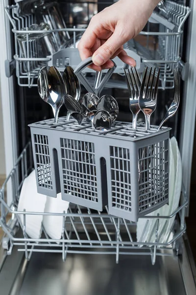 a woman\'s hand loads a basket with forks and spoons into the dishwasher. Washing dishes and maintaining the kitchen.