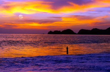 Günbatımında okyanusta bir adam dua ediyor. Akşam gökyüzünde ay yükselirken.
