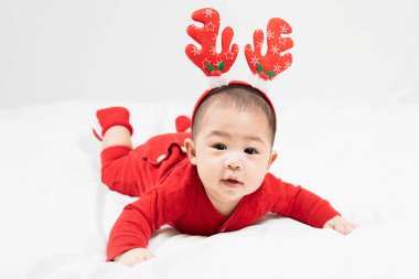 Yeni yıl ve Noel konsepti. 5 aylık Asyalı bebek geyik boynuzu takıyor. Evde yerde kırmızı elbiseler var. Tatil sezonu.
