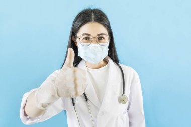 Tıbbi maske, eldiven, steteskop takan Asyalı genç bir kadın izole edilmiş mavi arka plan, onaylayan yüz ifadesini onaylıyor ve kameraya başarıyla bakıyor..