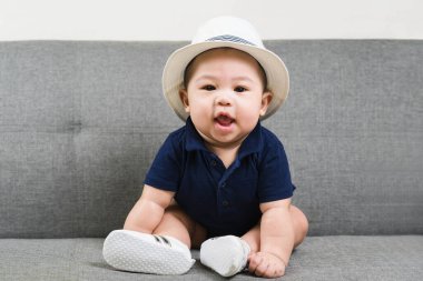 Kanepede ilk oturan sevimli erkek bebek. 5-6 aylık tatlı Asyalı bebek mavi tişört, beyaz şapka ve ayakkabılar giyiyor gülümsüyor ve kameraya bakıyor..