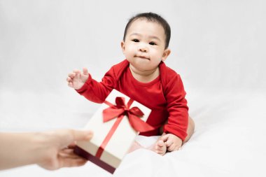 Kırmızı elbiseli şirin Asyalı bebek evde beyaz battaniyeye oturmuş hediye kutusunu alıyor. Yılbaşı hediyesini çocuğa, çocuğa, bebeğe, yeni yıla, yeni yıla, bebeklere, çocukluğa. seçici bir şekilde odaklanıyor..